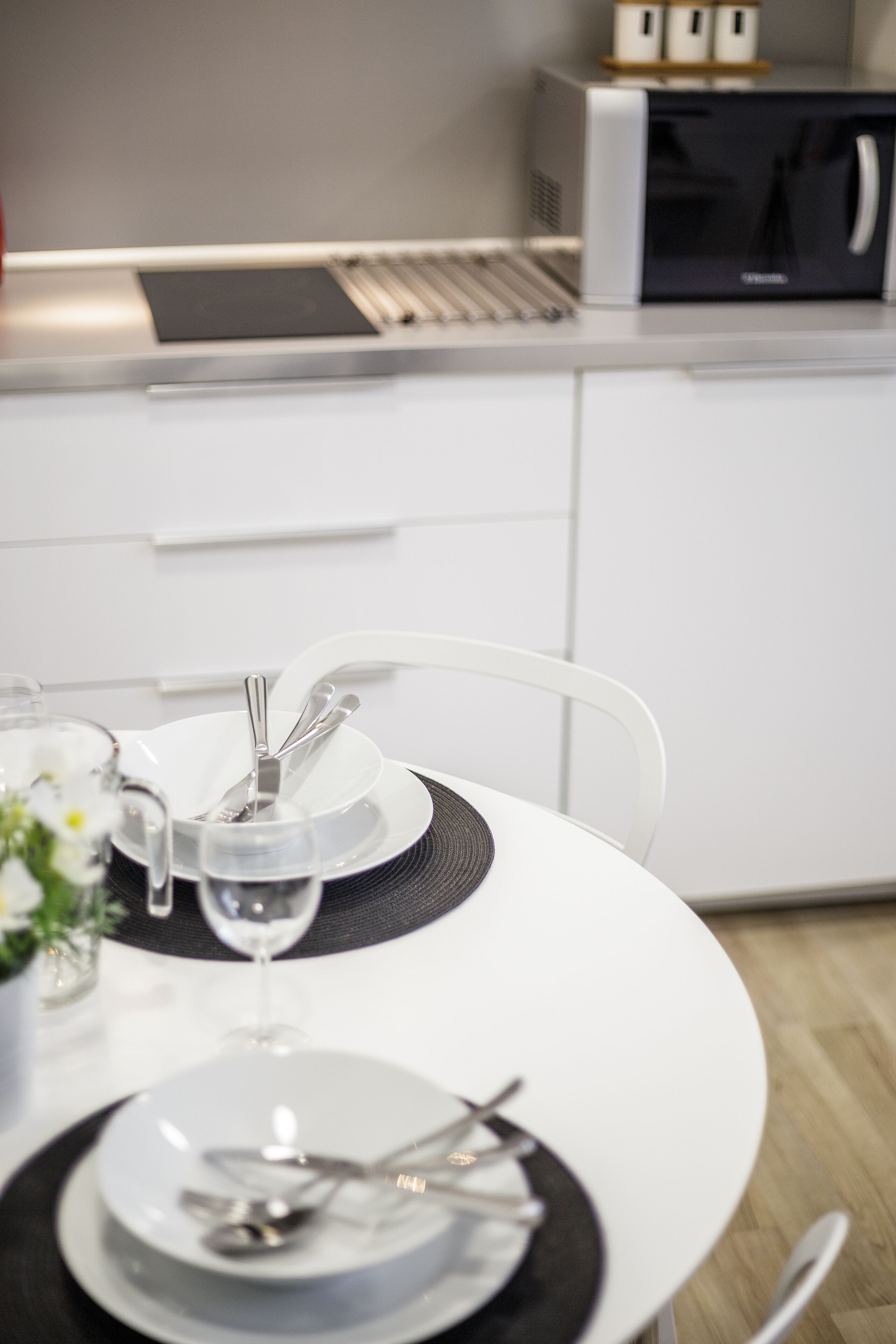 Una cocina moderna con armarios blancos, un microondas y un comedor con una mesa blanca dispuesta para dos personas.