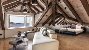 Een ruime slaapkamer onder de dakrand met een groot raam met uitzicht op zee, een moderne bank en een bed met hoofdeinde.