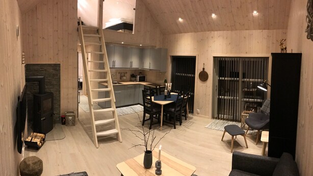 A modern interior with a wooden floor, a kitchen area, a dining table with chairs, a sofa, a ladder, and a fireplace. - Fam friendly furniture cottage in a quiet environment. 8 Pers Sauna and Jacuzzi (Stord)