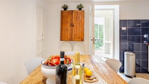 Een eethoek met een houten tafel, stoelen, een schaal tomaten, een fles wijn en een fles olijfolie.