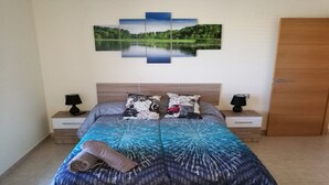 A bedroom with a bed, two bedside tables, a nightstand, a wooden door, and a wall-mounted artwork of a lake. - "A JUMP TO PARADISE ........ HOLIDAYS ALL YEAR ROUND". (SAN JORGE)