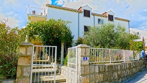 Edificio blanco con muro de piedra, portón metálico, letrero con el número 3 y abundante follaje.