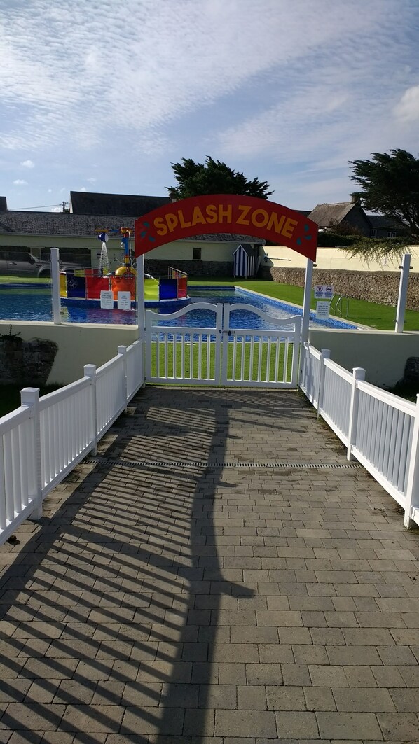 Ingresso alla zona splash con un arco rosso segnaletico, una piscina e una recinzione bianca.