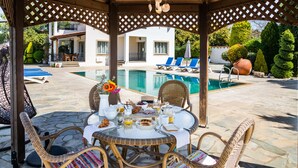 A covered outdoor dining area with wicker furniture, a table set with breakfast items, and a view of a swimming pool and a white building in the background. - VILLA DEMETRA (4 ensuite bedrooms,  stunning view,BBQ,HighSpeed WI-FI,swim.pool) (Argaka)