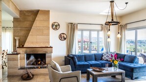 A living room with a fireplace, a blue sofa, a coffee table, and a view of the outdoors. - VILLA DEMETRA (4 ensuite bedrooms,  stunning view,BBQ,HighSpeed WI-FI,swim.pool) (Argaka)