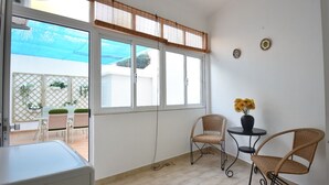 Una habitación con un ventanal grande, sillas de mimbre, una mesita con un florero y un armario blanco.