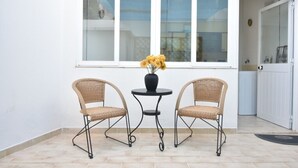 Un pequeño espacio al aire libre con dos sillas de mimbre y una mesa, un florero con flores, y una puerta blanca al fondo.