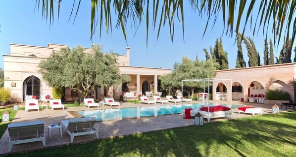 A poolside area with lounge chairs and a covered seating area, surrounded by greenery and a building with arches.