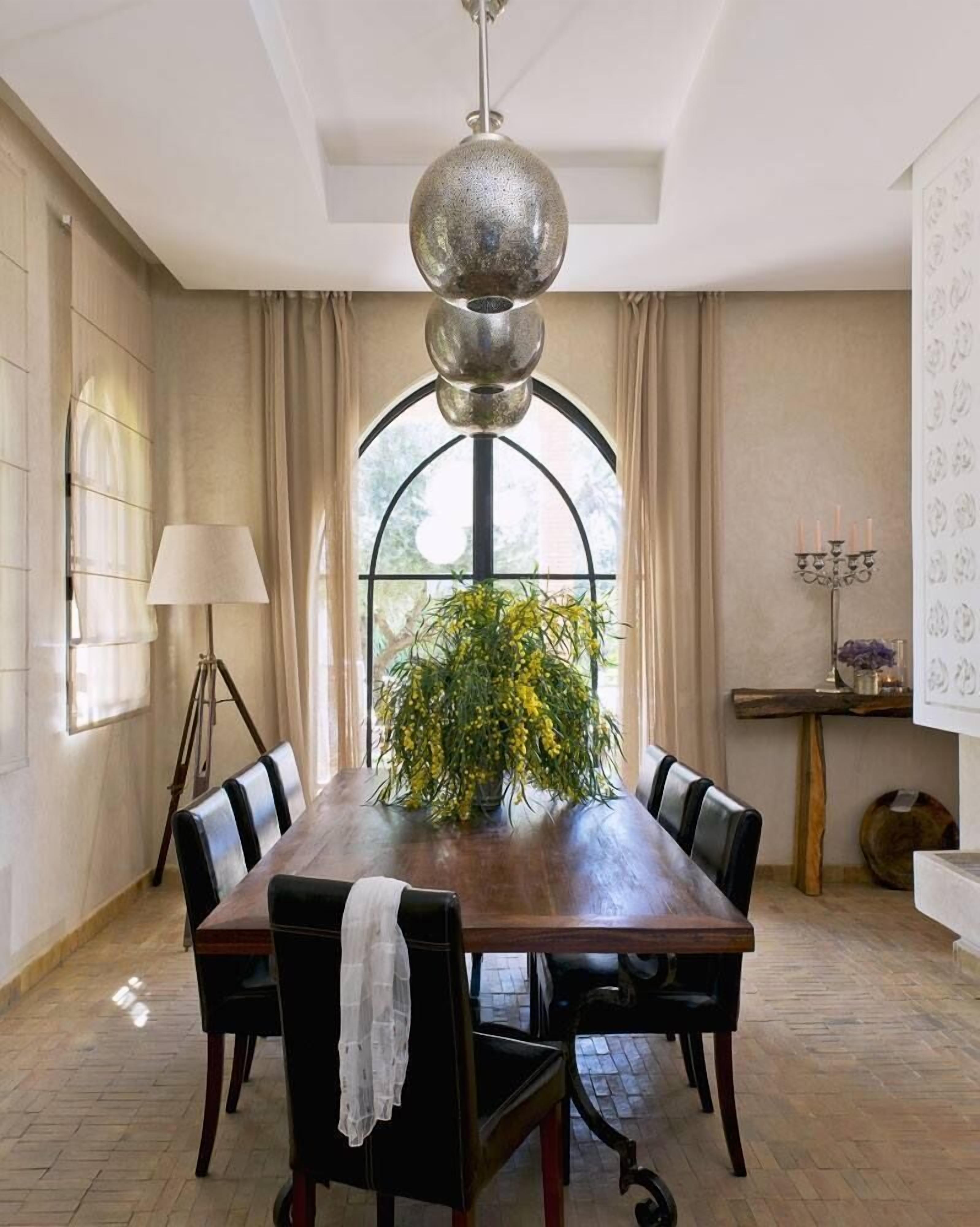 A dining room with a wooden table, six chairs, a chandelier, a window with curtains, and a vase with yellow flowers.