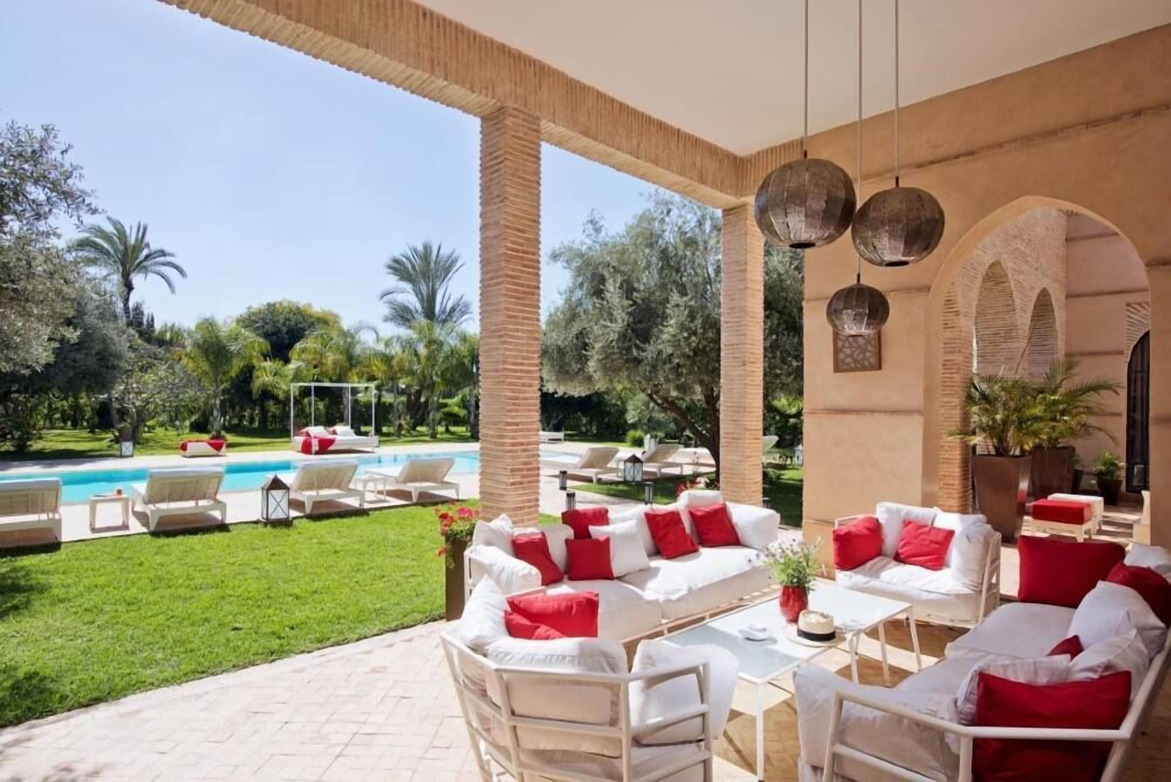 Un área de descanso al aire libre con mobiliario blanco y cojines rojos, una piscina y abundante vegetación.