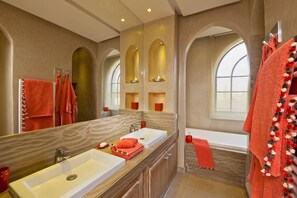 A bathroom with two sinks, a bathtub, and a large mirror. There are towels hanging on the wall and a window with blinds.