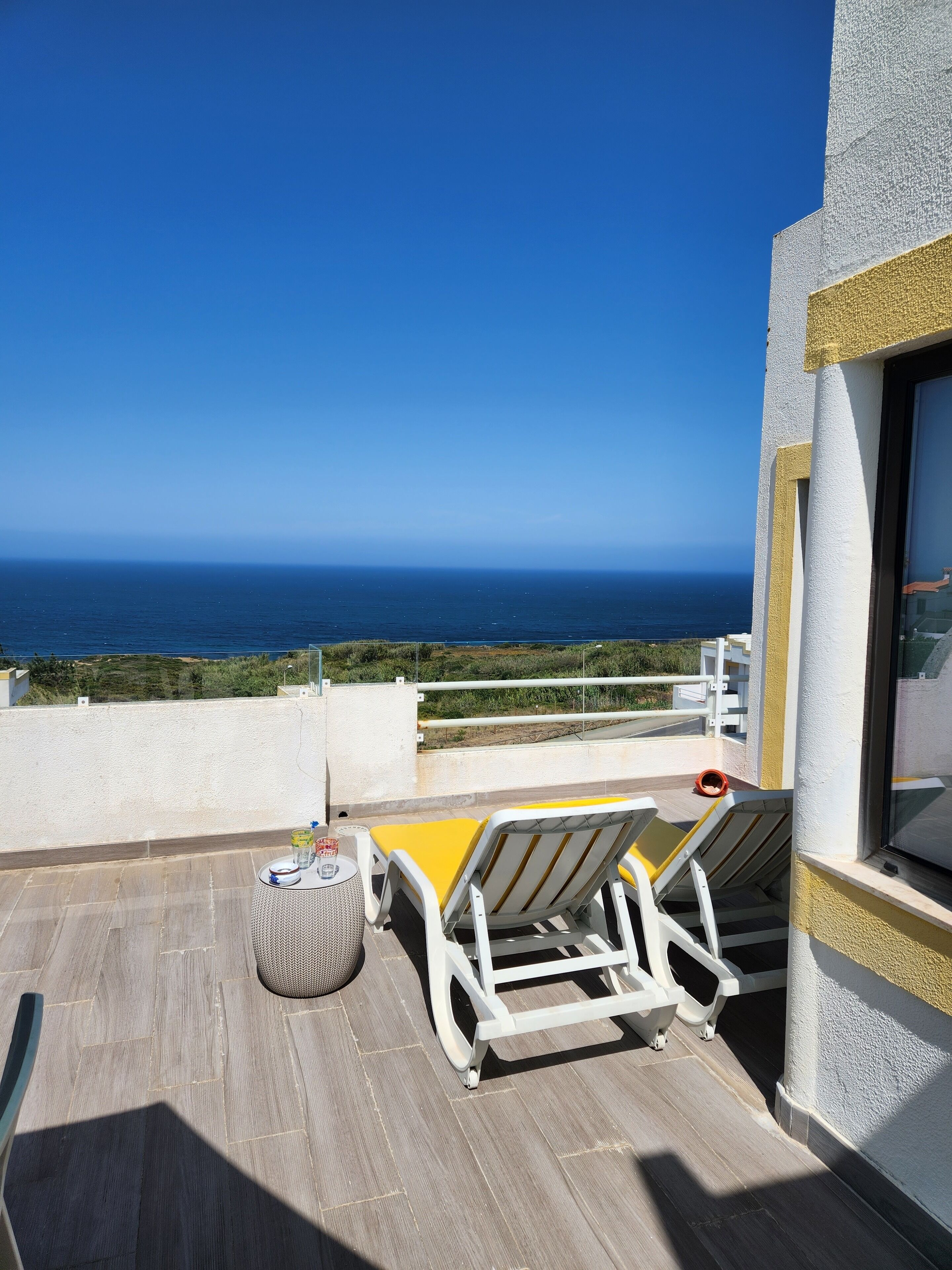 En balkon med udsigt over havet, en gul liggestol, et lille rundt bord og en flaske.