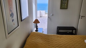 A bedroom with a bed, a lamp, a framed picture, and a view of the sea through an open door.