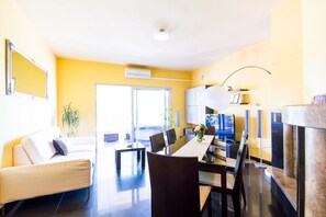 A modern living room with a sofa, dining table, and chairs. There is a large window, a plant, and a fireplace.
