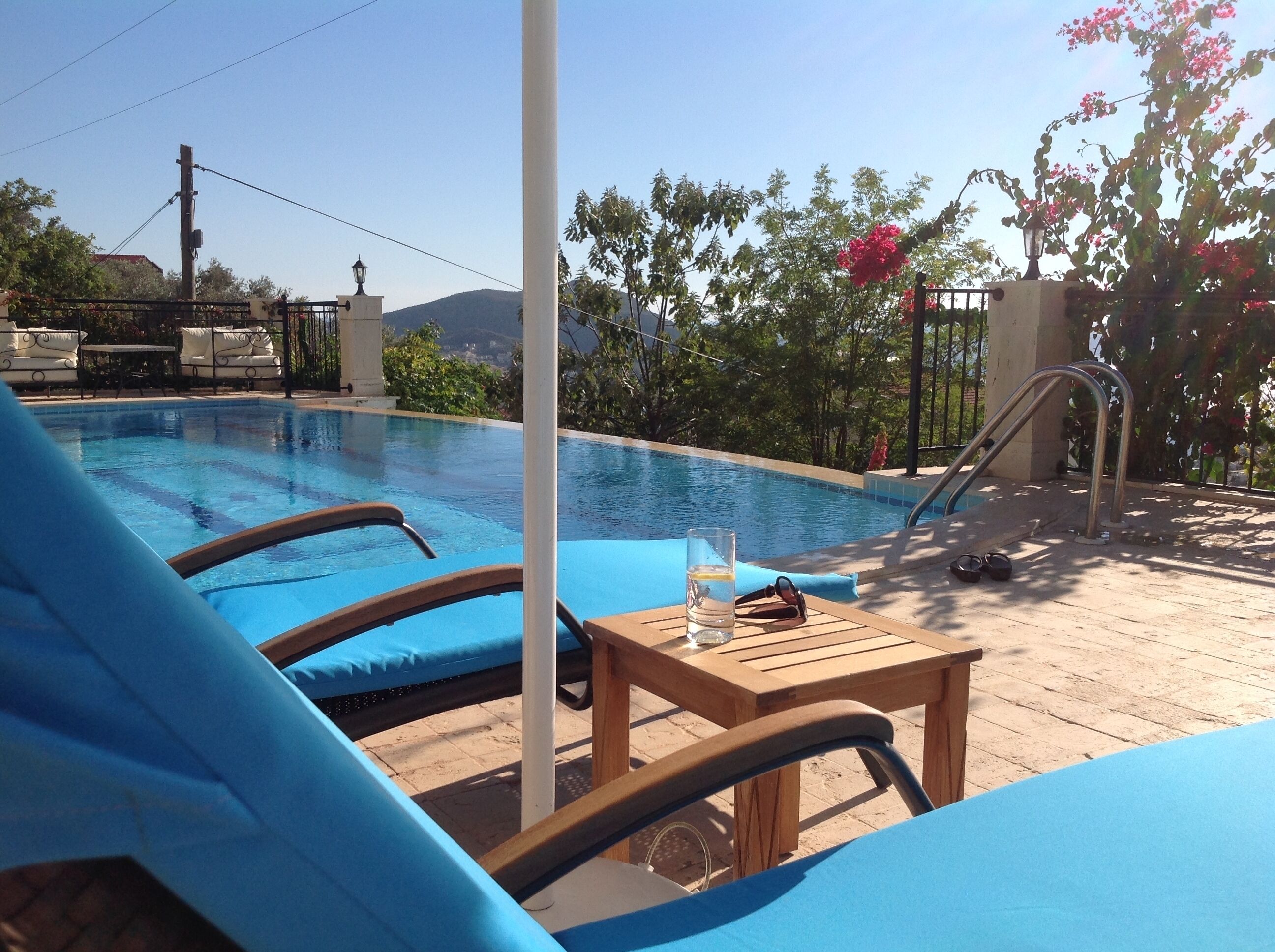 A pool area with lounge chairs and a table, surrounded by greenery and a view of hills.