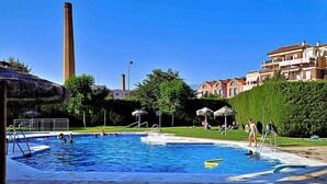 A swimming pool with a clear view of a tall chimney in the background, surrounded by lush greenery and residential buildings. - Spacious quiet apartment 450 meters to the beach, huge patio, communal pool. (Torre del Mar)