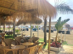An outdoor seating area with wicker furniture and thatched umbrellas, overlooking a beach with umbrellas and lounge chairs. - Spacious quiet apartment 450 meters to the beach, huge patio, communal pool. (Torre del Mar)