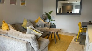 Ein modernes Wohnzimmer mit einem grauen Sofa, gelben und gemusterten Kissen, einem Holztisch und einem gelben Sessel. An der Wand hängt ein großer Spiegel.