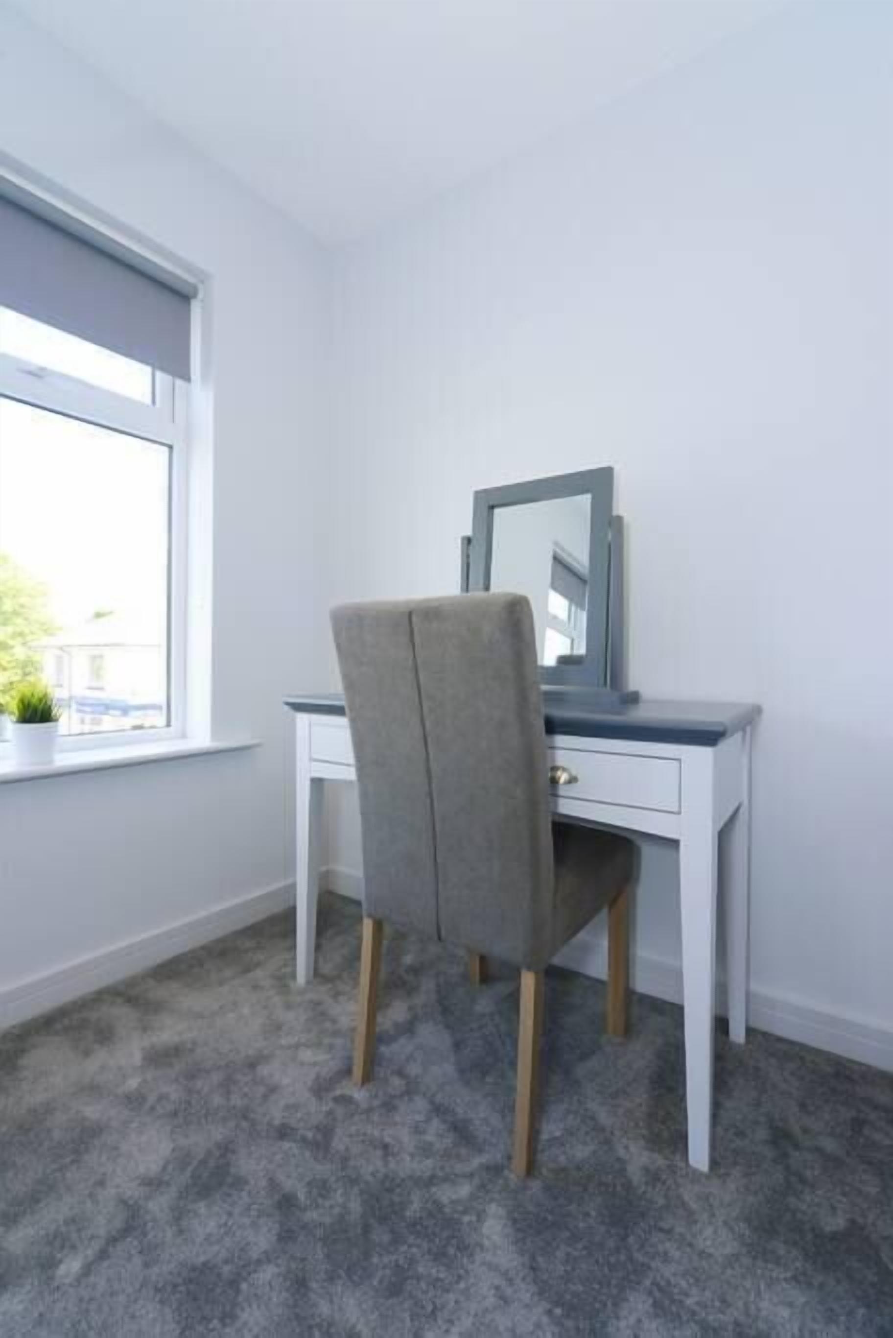A room with a grey upholstered chair, a white desk with a mirror, and a window with a view of buildings.