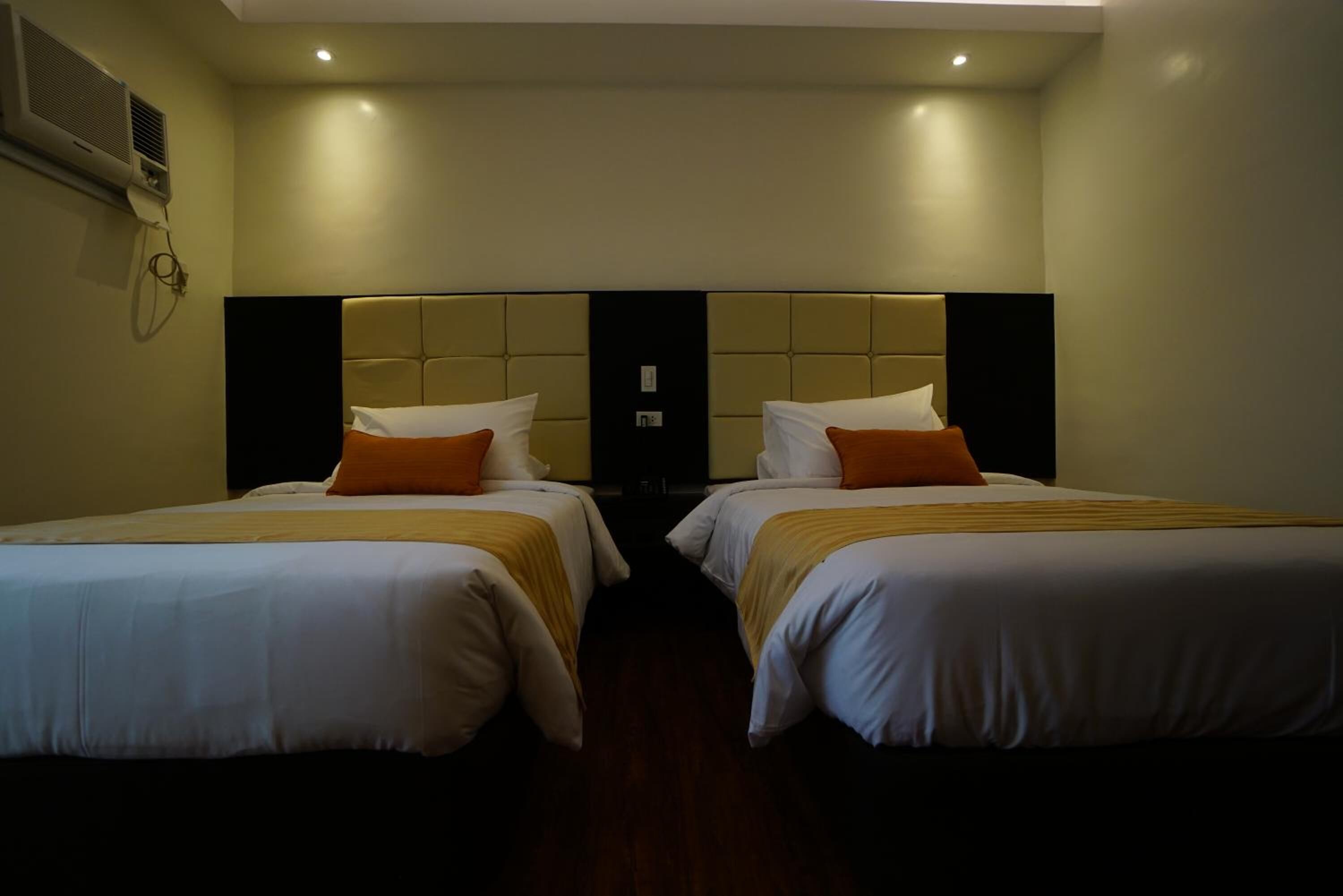 A hotel room with two beds, each with white bedding and orange pillows, a large headboard, and a wall-mounted air conditioning unit.