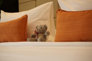 Una cama con ropa blanca y dos almohadas naranjas, un oso de peluche con un lazo rojo, y una almohada blanca al fondo.