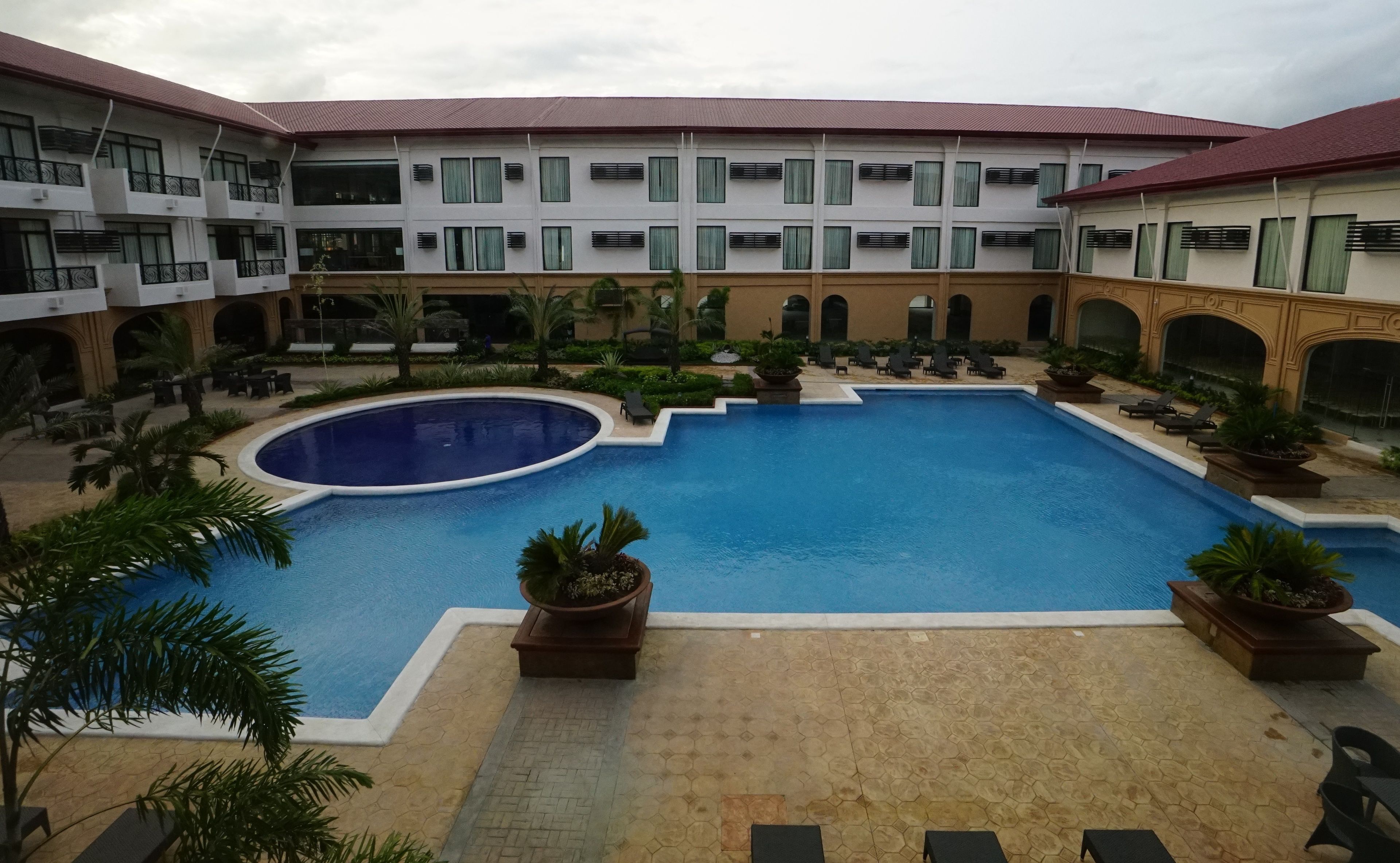 A hotel courtyard with a central swimming pool, multiple lounge areas, and a multi-story building with balconies.