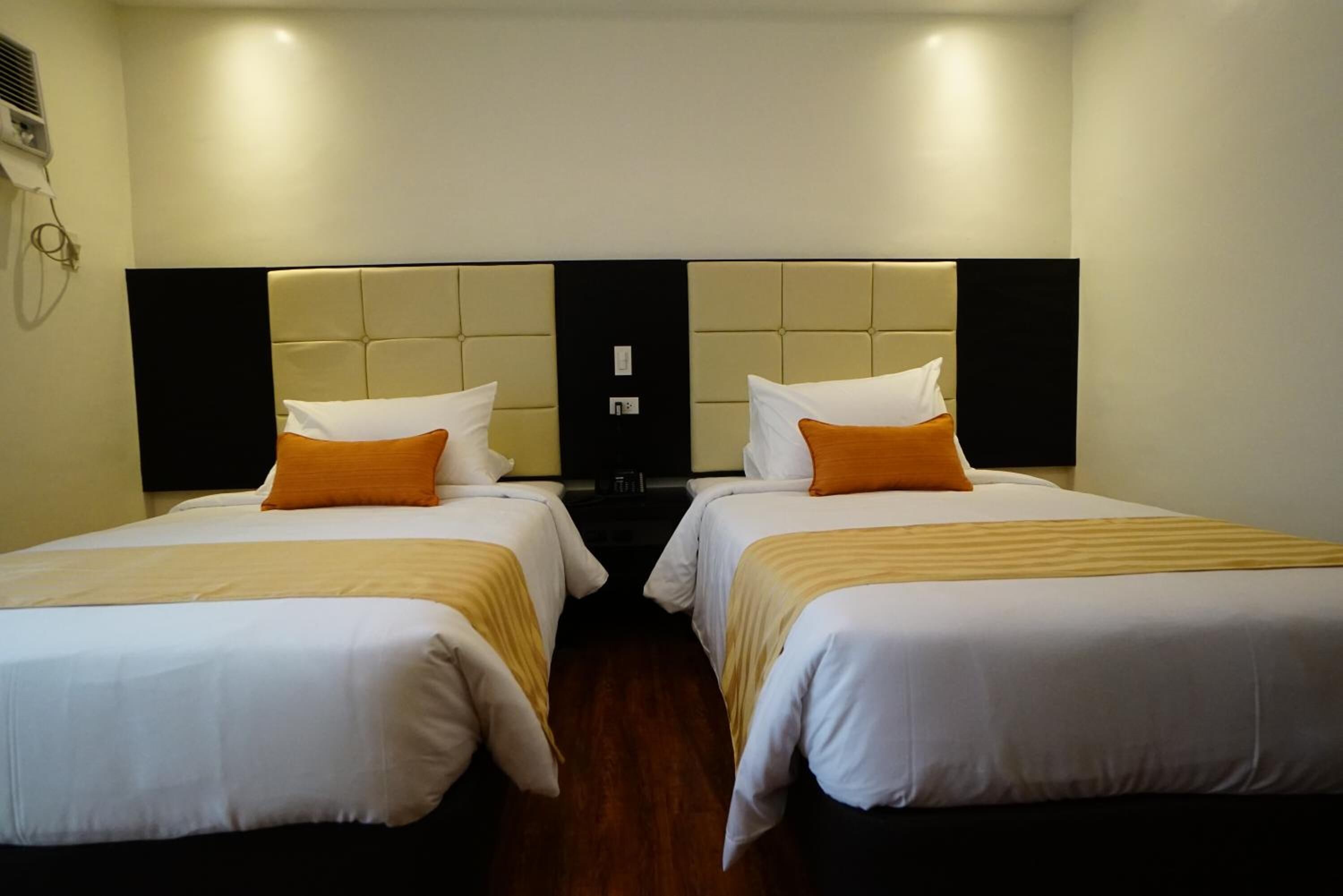 A hotel room with two beds, each with white bedding and orange pillows. The room has a modern design with a dark headboard and a wooden floor.