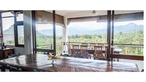 A dining area with a wooden table and chairs, a banana centerpiece, and a balcony with a view of mountains and greenery.