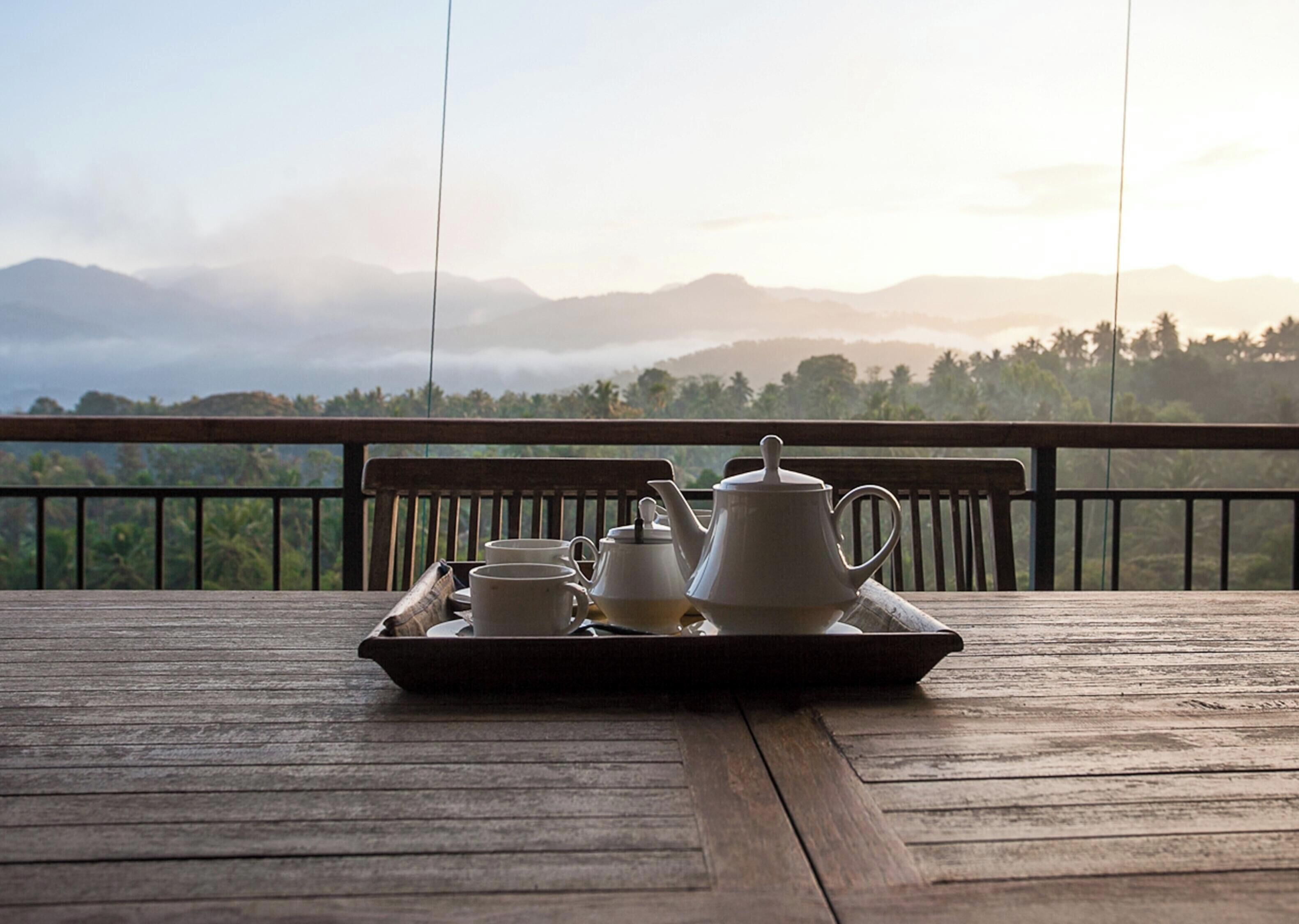 Una mesa de madera con una bandeja que sostiene una tetera, tazas y platillos, con montañas y un cielo nublado de fondo.