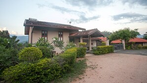 A two-story house with a tiled roof, surrounded by lush greenery and a gravel driveway.