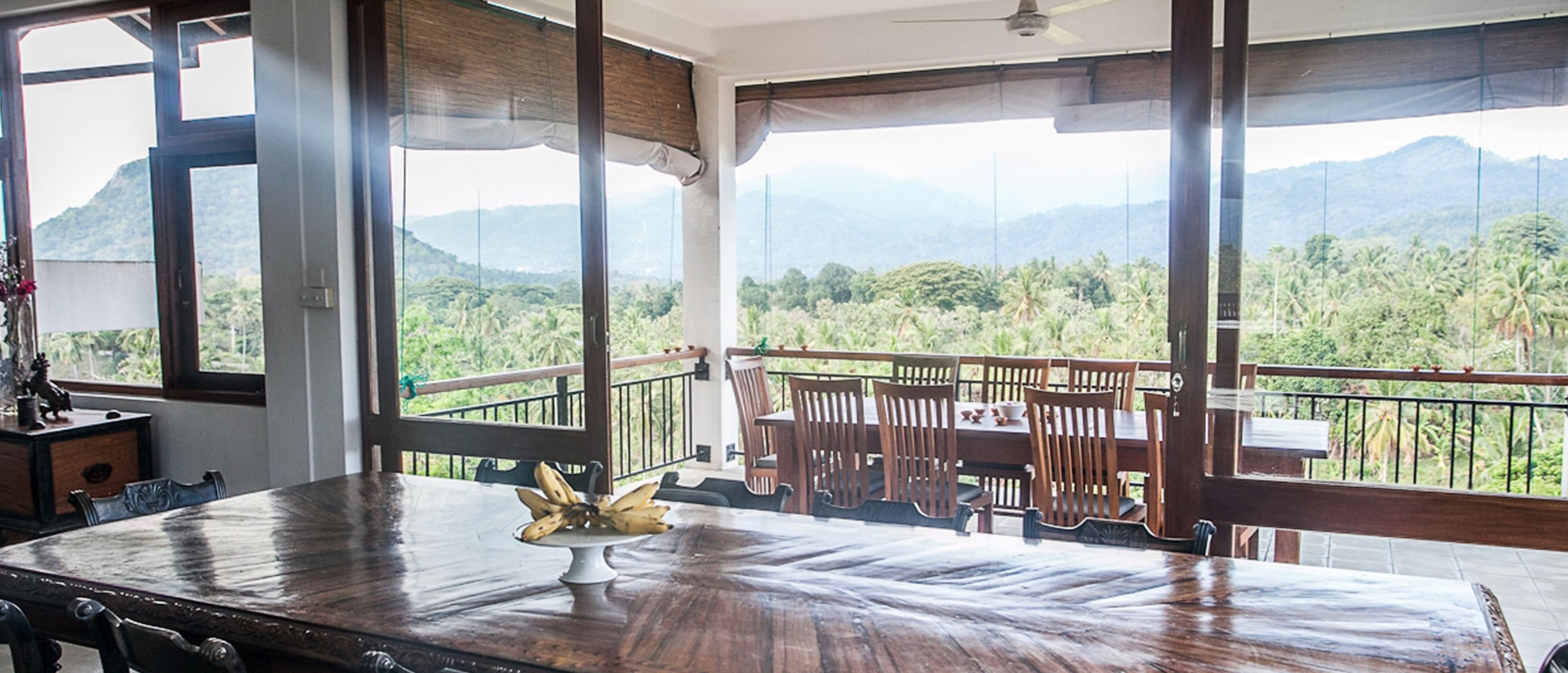 A dining area with a wooden table and chairs, a banana centerpiece, and a balcony with a view of mountains and greenery.