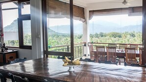 A dining area with a wooden table and chairs, a banana centerpiece, and a balcony with a view of mountains and greenery. - Tranquility in a golf and country resort- Luxury villa with live-in cook & staff (Rajawella)