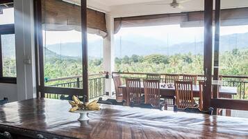 A dining area with a wooden table and chairs, a banana centerpiece, and a balcony with a view of mountains and greenery.