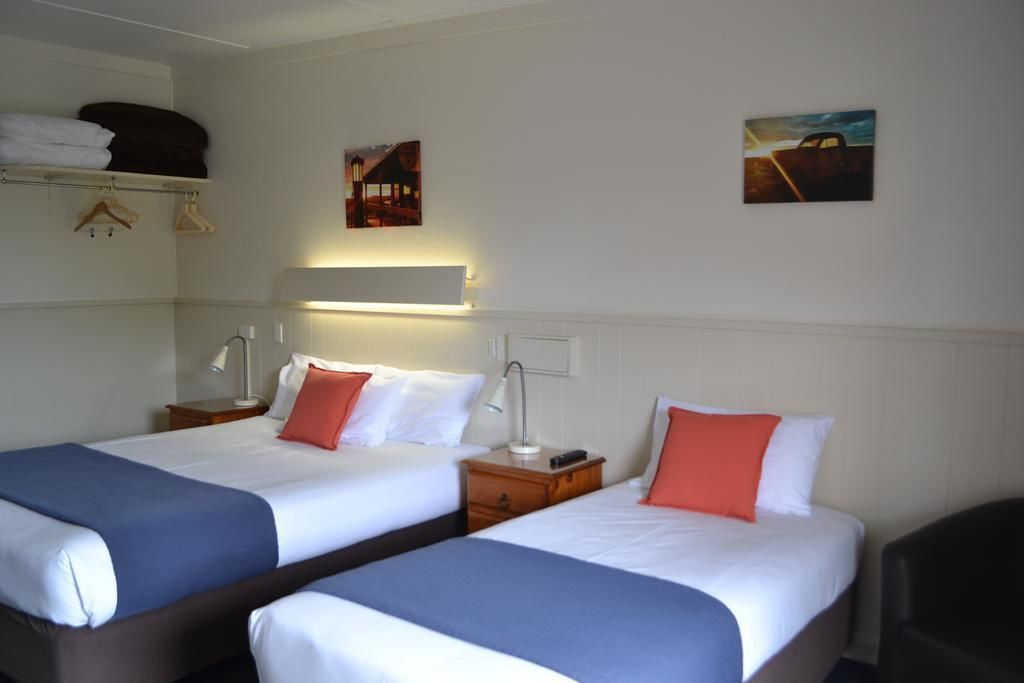 A hotel room with two beds, each with white bedding and blue headboards, a bedside table with a lamp, and a wall-mounted shelf above the beds.