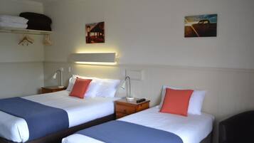 A hotel room with two beds, each with white bedding and blue headboards, a bedside table with a lamp, and a wall-mounted shelf above the beds.