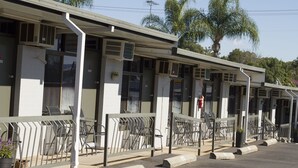 A row of motel rooms with individual balconies, air conditioning units, and a parking area with a palm tree in the background.