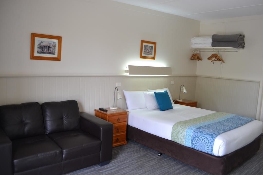 A hotel room with a bed, a leather sofa, a bedside table, a lamp, and a wall-mounted shelf with towels.