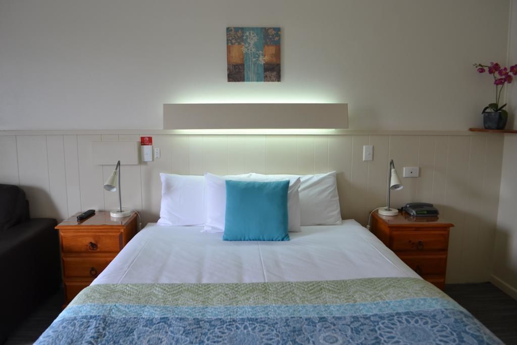 A hotel room with a bed, two bedside tables, a nightstand, a lamp, a painting on the wall, and a small potted plant.