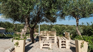 A stone-paved outdoor seating area with wooden benches and planters, surrounded by olive trees and greenery.