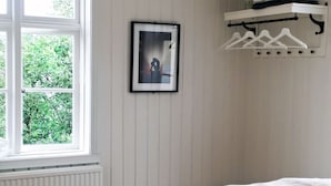 Et soveværelse med en stor seng, et vindue med persienner, en hængende plante og et billede i ramme på væggen.