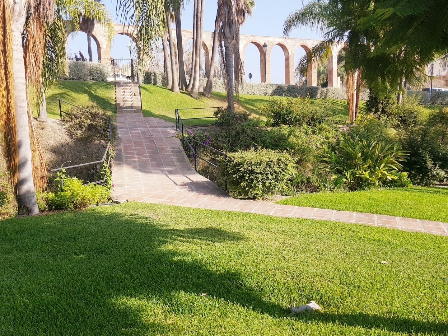 A pathway leading to a bridge with arches, surrounded by well-maintained grass and palm trees.