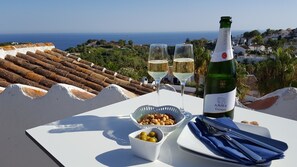 Ein Tisch mit einer Weinflasche, zwei Gläsern, einer Nussmischung, einer Oliven-Schale und einem gefalteten Serviettenbild auf einer Terrasse mit Blick auf eine Küstenlandschaft.