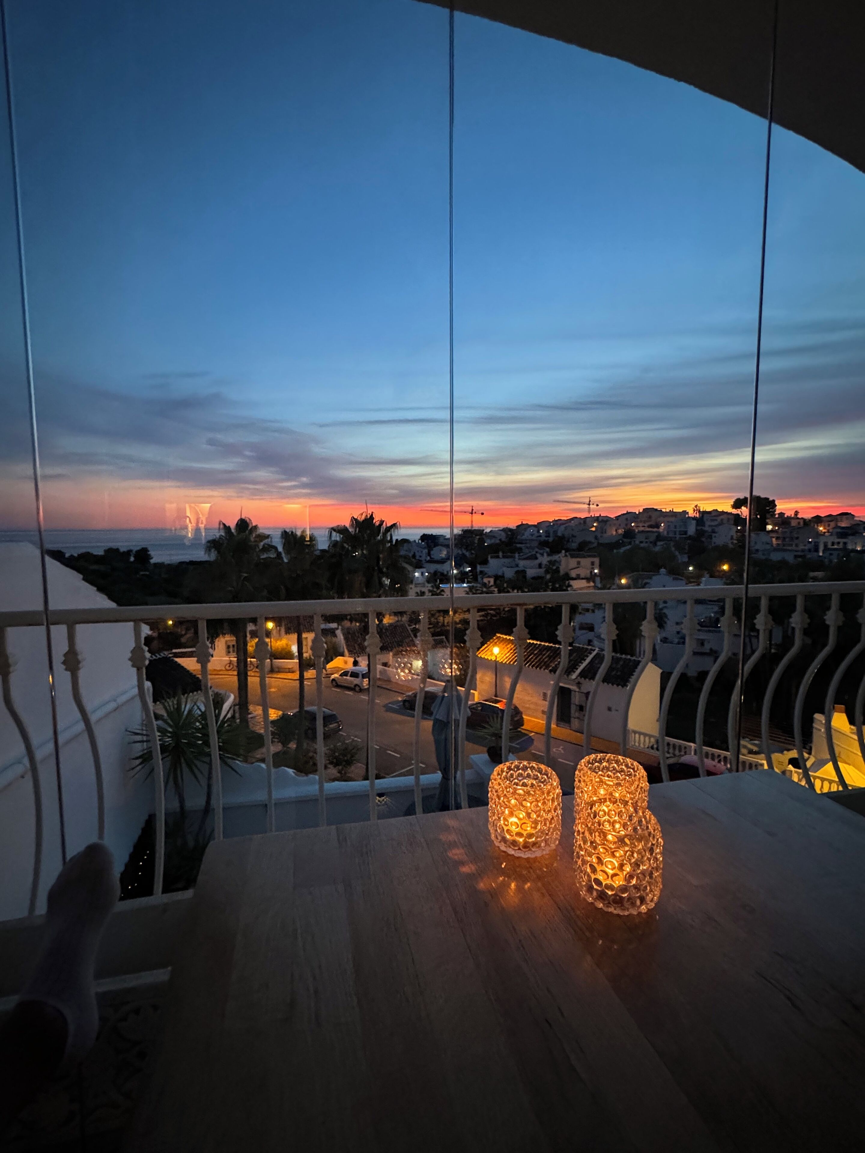 A balcony with a view of a sunset over a cityscape, featuring two illuminated candle-like lights on the table.