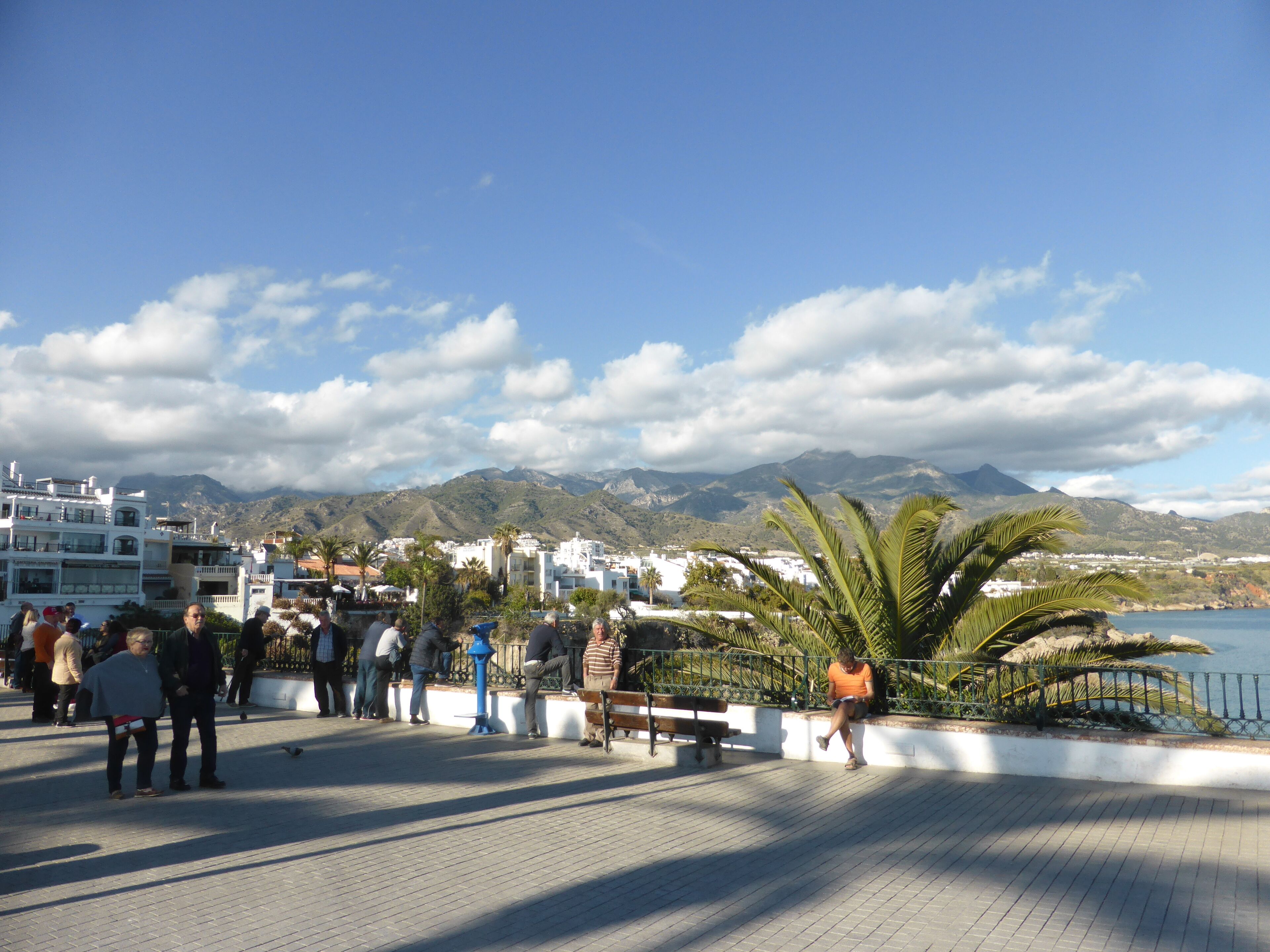 A public square with people, benches, and a view of mountains and water.