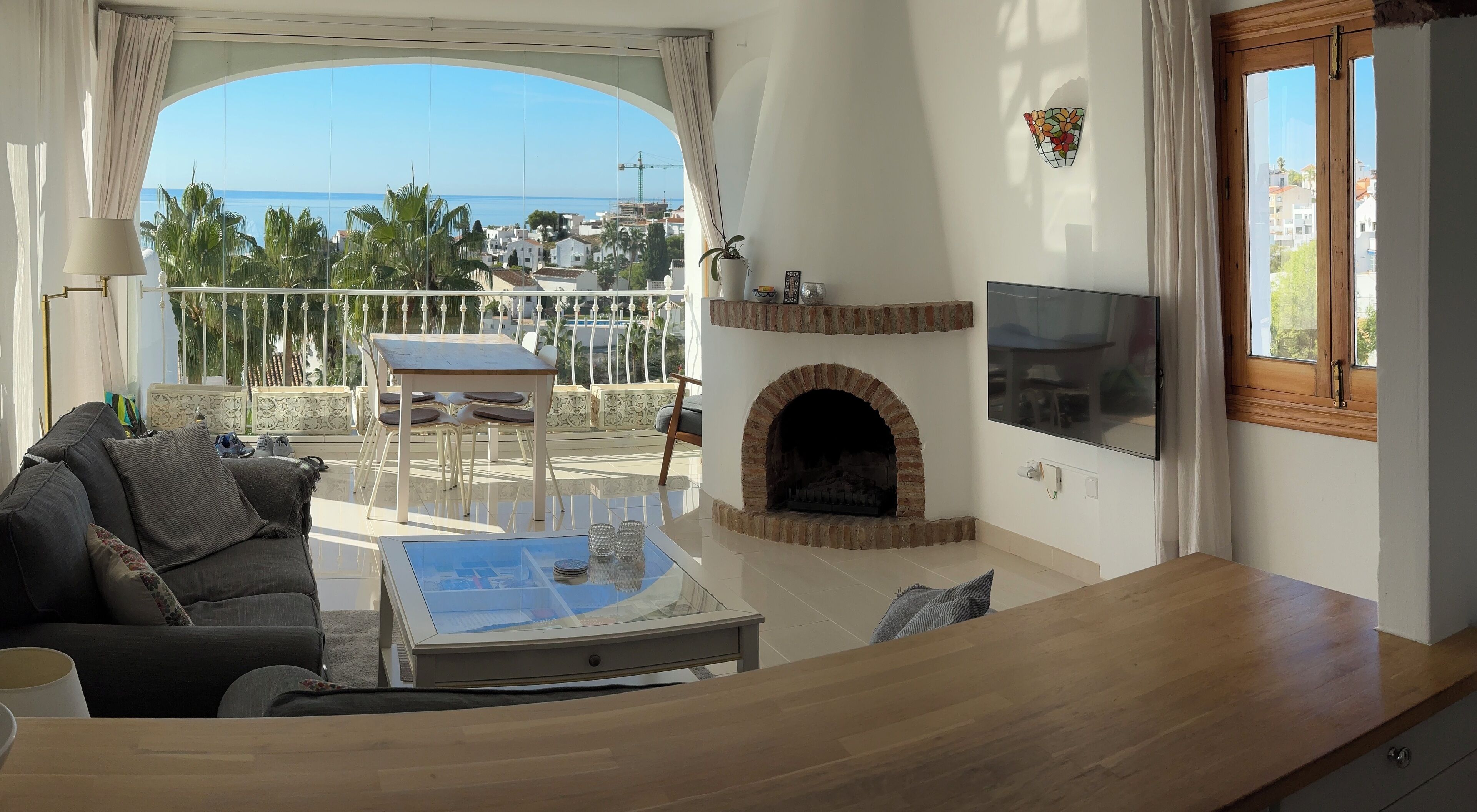 Un salon avec un canapé, une table et une cheminée. Il y a un balcon avec vue sur la mer et les bâtiments.