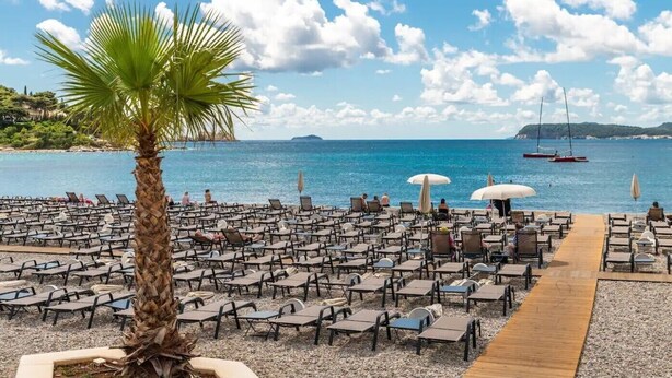 A beachside area with rows of sun loungers and umbrellas, a palm tree, and a view of the sea with boats. - 2 bedroom, 2 bathroom Apartment in Lapad with a large terrace and parking (Dubrovnik)