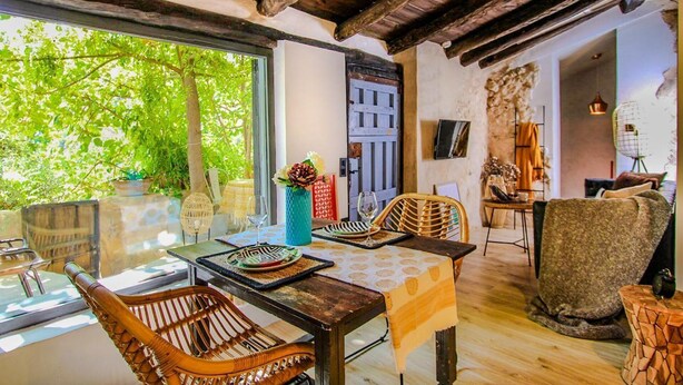 A room with a wooden ceiling, a dining table with chairs, a sofa, and a view of greenery outside. - HOHLE Cave Suite (Cave Hotel Lomas de Ruvira) (Jorquera)