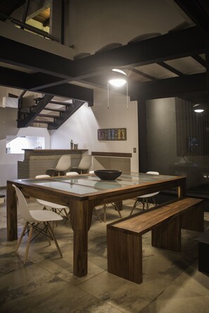 A modern kitchen with a large wooden table, white chairs, and a bench. There is a staircase in the background and a wall-mounted light fixture.