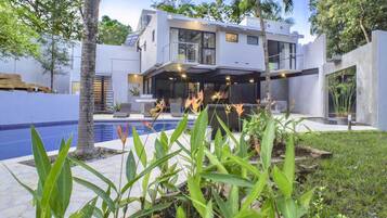 Une maison moderne à deux étages avec une piscine, entourée de plantes tropicales et de palmiers.