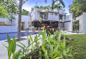 Una casa moderna de dos pisos con piscina, rodeada de plantas tropicales y palmeras.